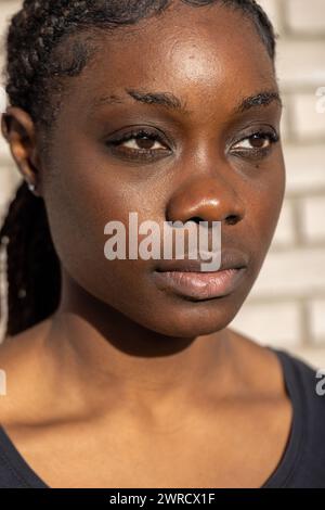 La photographie est un portrait rapproché d’une jeune femme africaine avec une expression réfléchie. Son regard est dirigé légèrement hors caméra, ajoutant une qualité franche à l'image. La lumière du soleil illumine un côté de son visage, créant un contraste dynamique avec le côté ombragé. Ses cheveux sont coiffés dans des tresses soignées qui encadrent son visage, et sa peau montre une belle lueur naturelle. La faible profondeur de champ garantit que les traits de la femme sont le point focal sur l'arrière-plan flou. Femme africaine ensoleillée avec cheveux tressés. Photo de haute qualité Banque D'Images