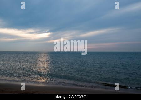 Cette image respire la tranquillité à travers un paysage marin serein où les derniers rayons du soleil traversent les nuages qui se séparent, projetant une douce lueur éthérée sur l'eau. La plage au premier plan est une étroite bande de sable, menant le regard du spectateur vers le vaste océan qui s'étend jusqu'à l'horizon. Les nuages sont rendus dans des tons de gris et de blanc, avec un soupçon de rose du soleil couchant, créant une atmosphère paisible mais sombre. Paysage marin tranquille avec les rayons du soleil perçant les nuages. Photo de haute qualité Banque D'Images