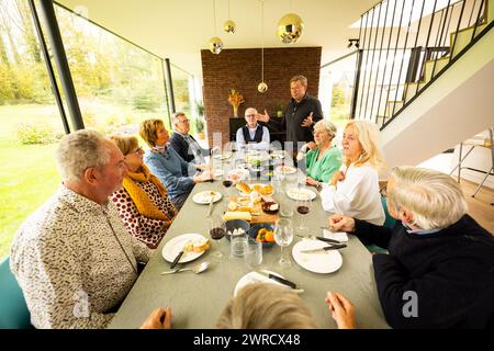 Un groupe animé d'adultes s'est réuni autour d'une table à manger élégante, partageant des histoires et des rires dans une chambre spacieuse et moderne avec vue sur le jardin. Famille et amis appréciant un repas convivial dans une élégante salle à manger vitrée. Photo de haute qualité Banque D'Images