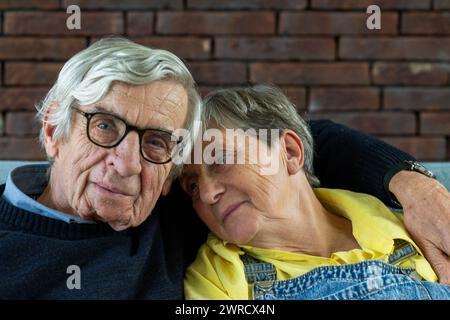 L'image représente un couple aîné affectueux s'appuyant l'un sur l'autre dans un moment d'intimité détendue. L'homme, avec des cheveux argentés et des lunettes, partage la monture avec une femme portant un haut jaune et une salopette en denim. Les deux semblent satisfaits et à l'aise l'un avec l'autre. La toile de fond d'un mur de briques ajoute une atmosphère rustique et accueillante à la scène, suggérant un cadre de chaleur et de familiarité. Couple senior détendu et proche ensemble. Photo de haute qualité Banque D'Images