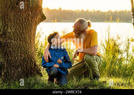 Cette image capture un moment tendre entre un grand-père et son petit-fils assis au bord d'un lac tranquille au coucher du soleil. L'homme âgé, avec une barbe blanche, des lunettes et un sourire chaleureux, place doucement sa main sur l'épaule du garçon, transmettant un sentiment de guidage et d'affection. Le jeune garçon le regarde avec un regard d'admiration et de confiance. Elles sont encadrées par un grand arbre, et le doux contre-jour du soleil couchant crée une atmosphère sereine et chaleureuse, soulignant ce moment intime en famille. Moment tendre entre grand-père et petit-fils par un lac. Photo de haute qualité Banque D'Images