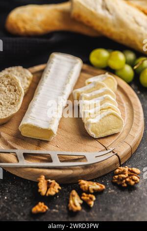 Coin de fromage Brie coiffé sur un plateau de fromage en bois accompagné de pain français frais et de raisins pour une configuration culinaire rustique Banque D'Images