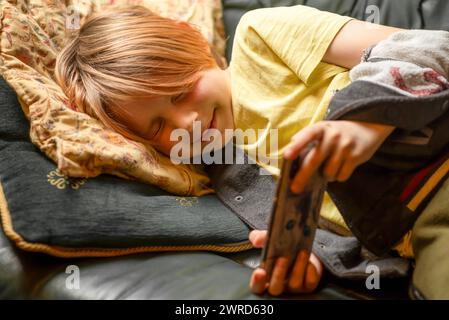 Un jeune garçon de 10 ans est allongé sur le canapé avec un téléphone dans les mains. Selon Internet, jeux téléphoniques Banque D'Images