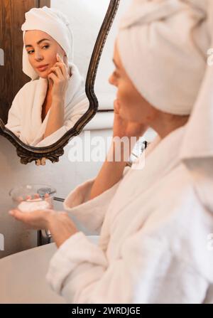 Femme attrayante applique la crème hydratante sur son visage portant un peignoir blanc et une serviette sur la tête debout près du miroir dans la salle de bain. Soins de la peau pour WO Banque D'Images