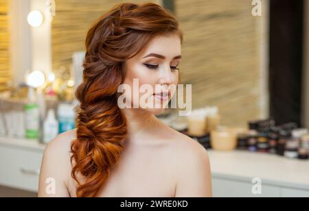 Belle fille, avec de longues cheveux roux poilus. Coiffeur tisse une tresse française, gros plan dans un salon de beauté. Soins capillaires professionnels et création de cheveux Banque D'Images