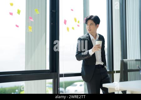 Homme d'affaires asiatique buvant un café et regardant par une fenêtre dans le bureau à domicile, homme d'affaires attrayant travaillant dans le poste de travail. Banque D'Images