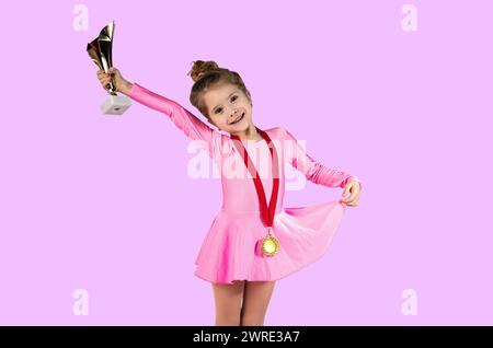 Portrait d'une petite fille gymnaste juste après avoir gagné vêtue d'une robe rose avec une médaille d'or autour de son cou, tenant le trophée nouvellement gagné dans son h. Banque D'Images