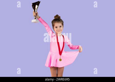 Portrait d'une petite fille gymnaste juste après avoir gagné vêtue d'une robe rose avec une médaille d'or autour de son cou, tenant le trophée nouvellement gagné dans son h. Banque D'Images