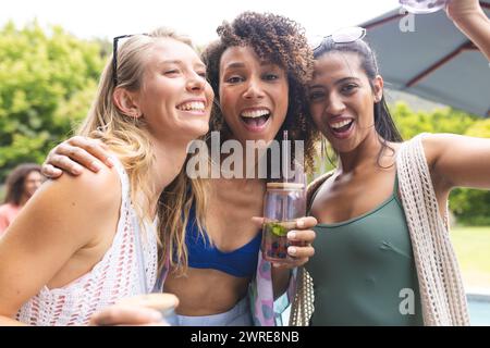 Trois jeunes amies féminines diverses sourient et posent de près pour un selfie, avec des boissons à la main Banque D'Images