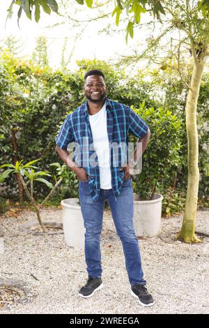 Homme afro-américain se tient souriant dans un jardin, portant une chemise à carreaux bleus et un Jean Banque D'Images