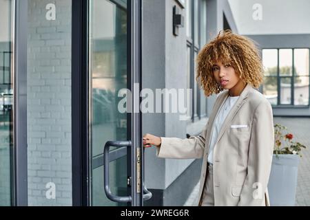 femme d'affaires afro-américaine bouclée en tenue formelle entrant dans l'immeuble de bureaux, jeune professionnel Banque D'Images