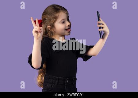 Photo de jolie petite fille charmante en t-shirt noir prenant selfie avec un appareil moderne montrant un fond violet isolé avec le signe V. Phot de haute qualité Banque D'Images