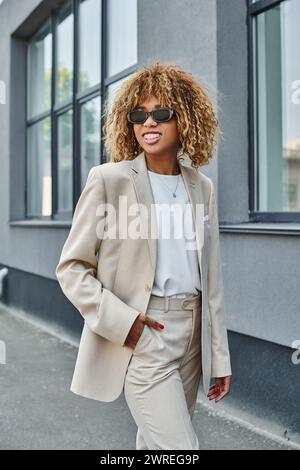 femme afro-américaine joyeuse avec des bretelles posant en costume et lunettes de soleil près de l'immeuble de bureaux Banque D'Images