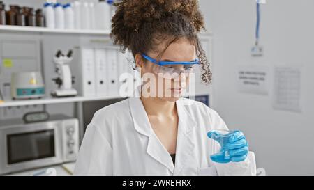 Une jeune femme hispanique aux cheveux bouclés portant des lunettes de sécurité et une blouse de laboratoire mène une expérience en laboratoire. Banque D'Images
