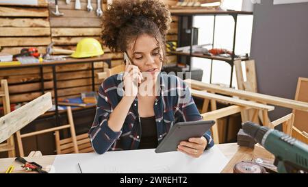 Une jeune femme hispanique aux cheveux bouclés est engagée dans une conversation téléphonique tout en tenant une tablette dans un atelier de menuiserie. Banque D'Images