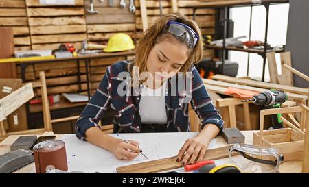 Séduisante jeune femme hispanique menuisière, habillée pour la sécurité avec des lunettes, note habilement des notes dans son atelier de menuiserie Banque D'Images