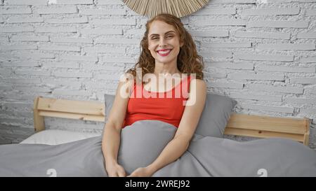 Jeune femme confiante, rayonnant de bonheur, est assise confortablement sur son lit, souriant largement dans sa chambre confortable, profitant de la tranquillité intérieure de son b Banque D'Images