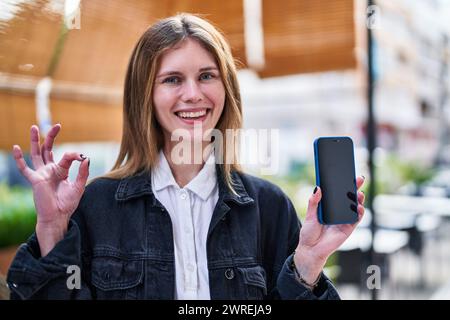 Jeune femme blonde souriante, debout sur une terrasse de café, tient son smartphone et donne le geste convivial OK-signe, symbolisant une excellente joie Banque D'Images
