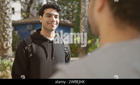 Deux hommes hispaniques souriant et discutant sur un sentier ensoleillé du parc, exsudant amitié et loisirs dans un cadre urbain en plein air. Banque D'Images