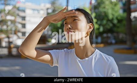 Une jeune femme asiatique protège ses yeux du soleil tout en se tenant dehors dans un parc de la ville. Banque D'Images