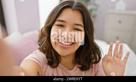 Une jeune femme hispanique joyeuse prend un selfie dans une chambre confortable, rayonnant de chaleur et de positivité. Banque D'Images