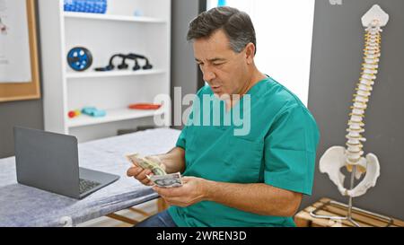 Médecin masculin mature dans les gommages examinant les dollars singapouriens dans le bureau de la clinique avec ordinateur portable et modèle de colonne vertébrale. Banque D'Images