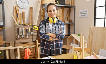 Femme mature confiante avec des protecteurs d'oreilles se tient les bras croisés dans un atelier de menuiserie lumineux. Banque D'Images