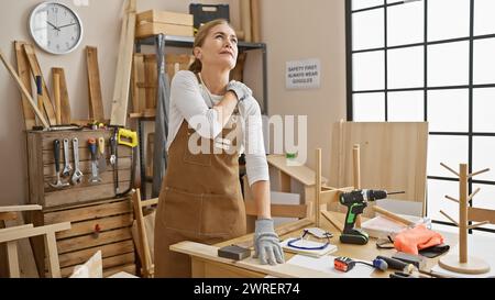Femme mature dans le tablier ressentant la douleur à l'épaule tout en travaillant dans un atelier de menuiserie avec des outils autour. Banque D'Images