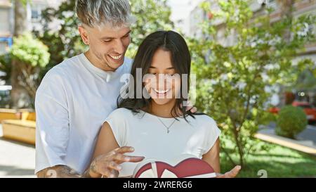 Un homme surprend une femme avec un cadeau en forme de cœur à l'extérieur, mettant en valeur l'amour, l'affection et une relation joyeuse. Banque D'Images