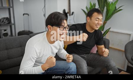 Deux hommes asiatiques célébrant avec enthousiasme tout en étant assis sur un canapé dans un environnement de salon confortable. Banque D'Images