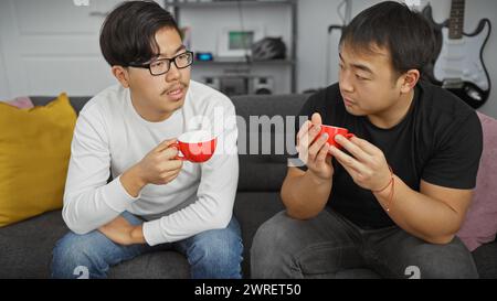 Deux hommes asiatiques ayant une conversation dans un salon moderne tout en tenant des tasses de café rouges Banque D'Images