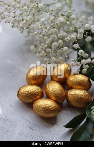 Œuf de Pâques en chocolat enveloppé dans une feuille d'or. Fleurs de printemps blanches et bonbons ester vue du dessus photo. Nature morte printanière brillante. Banque D'Images