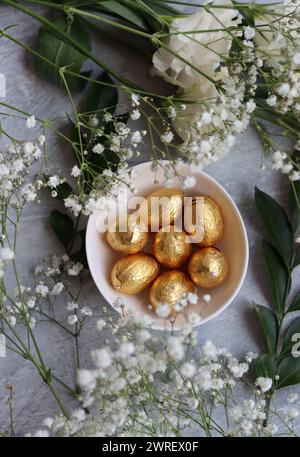 Œuf de Pâques en chocolat enveloppé dans une feuille d'or. Fleurs de printemps blanches et bonbons ester vue du dessus photo. Nature morte printanière brillante. Banque D'Images