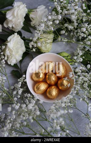 Œuf de Pâques en chocolat enveloppé dans une feuille d'or. Fleurs de printemps blanches et bonbons ester vue du dessus photo. Nature morte printanière brillante. Banque D'Images