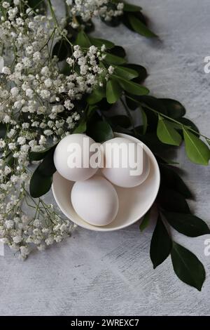 Composition simple nature morte avec des œufs et des fleurs blanches. Concept de célébration de Pâques. Oeufs dans un bol en céramique blanche vue de dessus photo. Banque D'Images