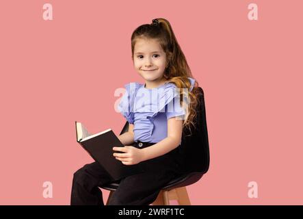 Portrait d'une petite fille souriante et attrayante tenant un livre et souriant à la caméra magnifiquement, dans un studio avec un fond rose. Haute qualité Banque D'Images