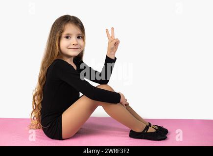 Vue latérale de la petite fille gymnaste regardant la caméra montrant le signe de paix portant un survêtement noir assis sur un tapis de yoga rose et un fond de studio blanc. Bonjour Banque D'Images