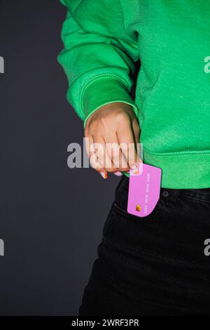 La photo d'une jeune femme sur un fond gris et avec un pull vert tenant une carte rose, la couleur aimée par les femmes. Elle le met dans sa poche Banque D'Images