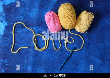 Mot Love fait de bande de roulement de laine jaune sur fond texturé bleu avec espace de copie. Photo vue de dessus de fil de laine et crochet. Passe-temps et loisirs Banque D'Images