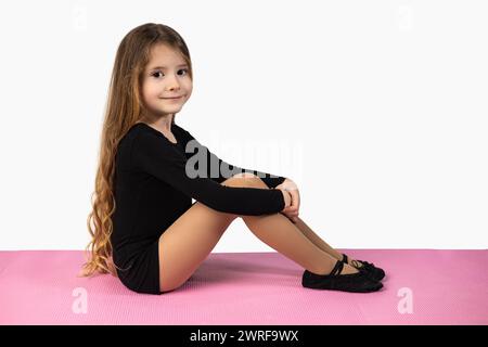 Vue latérale de la petite fille gymnaste regardant la caméra portant un survêtement noir assis sur un tapis de yoga rose et un fond de studio blanc. Photo de haute qualité Banque D'Images