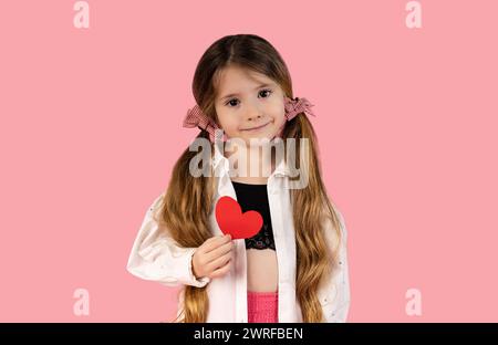 Une petite fille avec deux queues de cochon attrapées avec deux sommets roses, qui est photographiée sur un fond rose tenant un cœur, est dédiée à sa mère ou à tous Banque D'Images