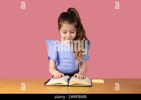 Image sur un fond rose avec une très belle petite fille avec deux tresses dans les cheveux, la petite fille est attrapée en lisant des livres dans le sch Banque D'Images