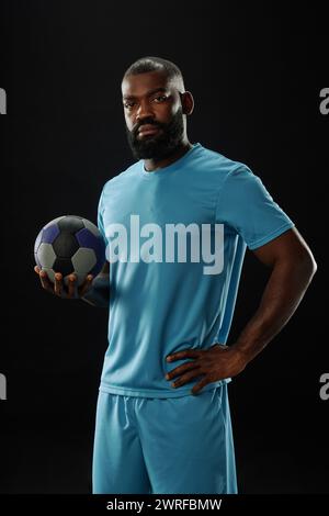 Portrait vertical de l'homme afro-américain confiant en tant que joueur de football regardant la caméra et portant l'uniforme de sport bleu debout sur fond noir Banque D'Images