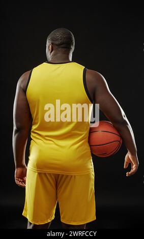 Vue arrière verticale d'un joueur de basket-ball musclé tenant le ballon tout en se tenant debout sur fond noir Banque D'Images
