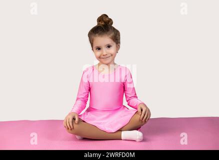 Petite fille blonde en robe rose assise sur un tapis de fitness avec ses jambes croisées et regardant la caméra avec un sourire sur un fond blanc. Qua. Élevé Banque D'Images