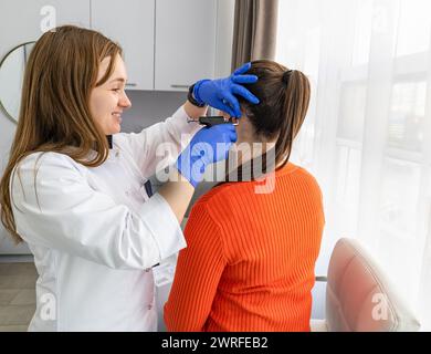 Vue de côté d'un médecin esthéticien portant des gants stériles bleus et un pistolet de perçage à la main prêt à percer l'oreille de la jeune brune pour appliquer la boucle d'oreille médicale. Hig Banque D'Images