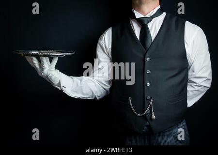 Maître d'hôtel ou serveur habillé avec gilet de costume noir et gants blancs tenant élégamment le plateau de service. Industrie des services et hospitalité professionnelle. Banque D'Images