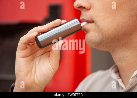 Un homme barbu fume des cigarettes électroniques grises sur fond rouge. Banque D'Images