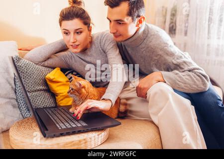 Couple de jeunes familles regardant la vidéo en ligne avec un chat utilisant un ordinateur portable à la maison.Un animal de compagnie au gingembre assis sur un canapé qui regarde l'écran.Curieux animal intéressé par f Banque D'Images