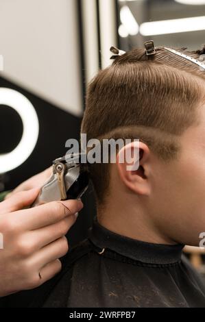 Barber rase le contour de la ligne ovale avec une tondeuse sur la tête du client. Un homme avec une barbe se fait couper les cheveux dans une chaise de salon de coiffure. Banque D'Images